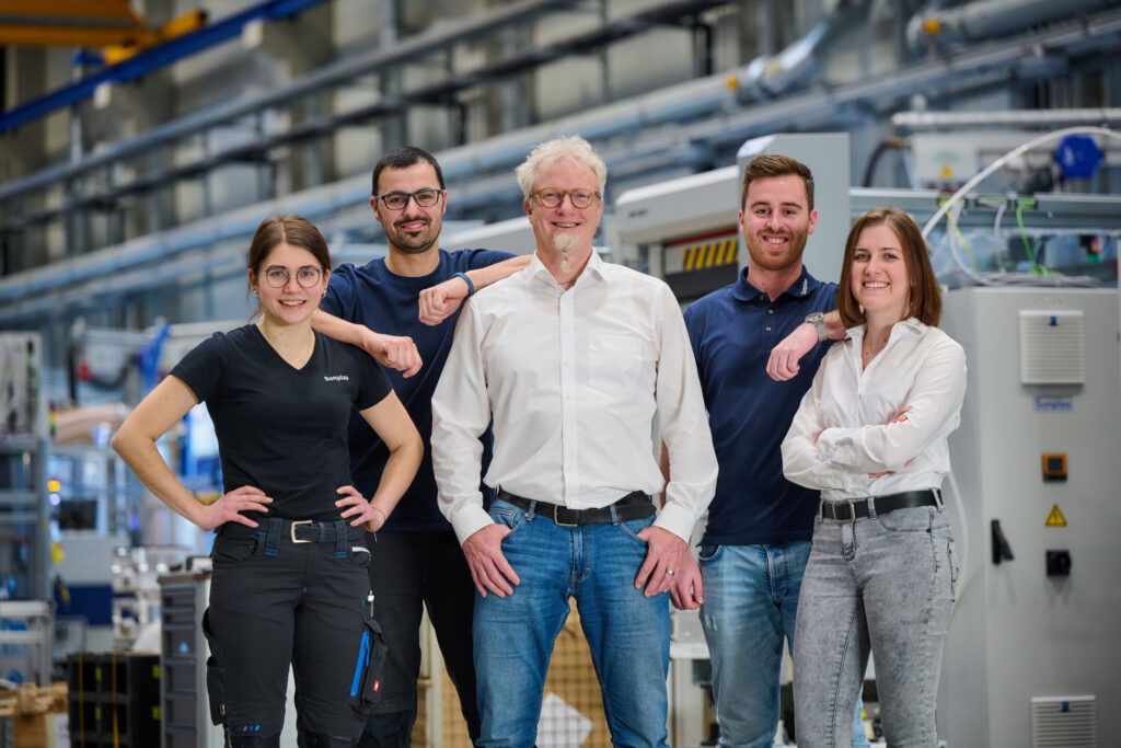 Fünf Personen stehen in der Montagehalle nebeneinander, schauen in die Kamera. Darunter sind zwei Frauen und drei Männer